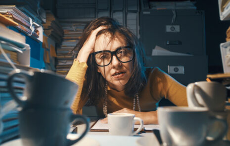 Overwhelmed woman with head on hand siting at desk surrounded by coffee mugs